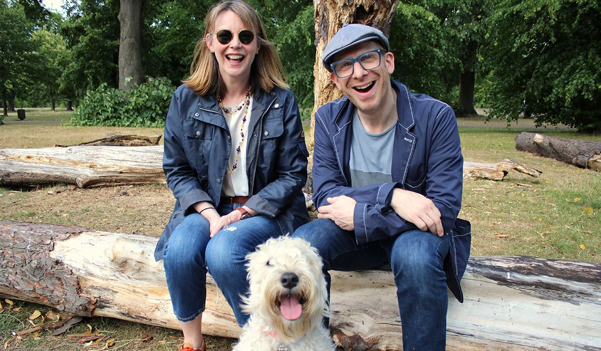 Two smiling people are sitting on a log in woodland with a white, curly-haired dog